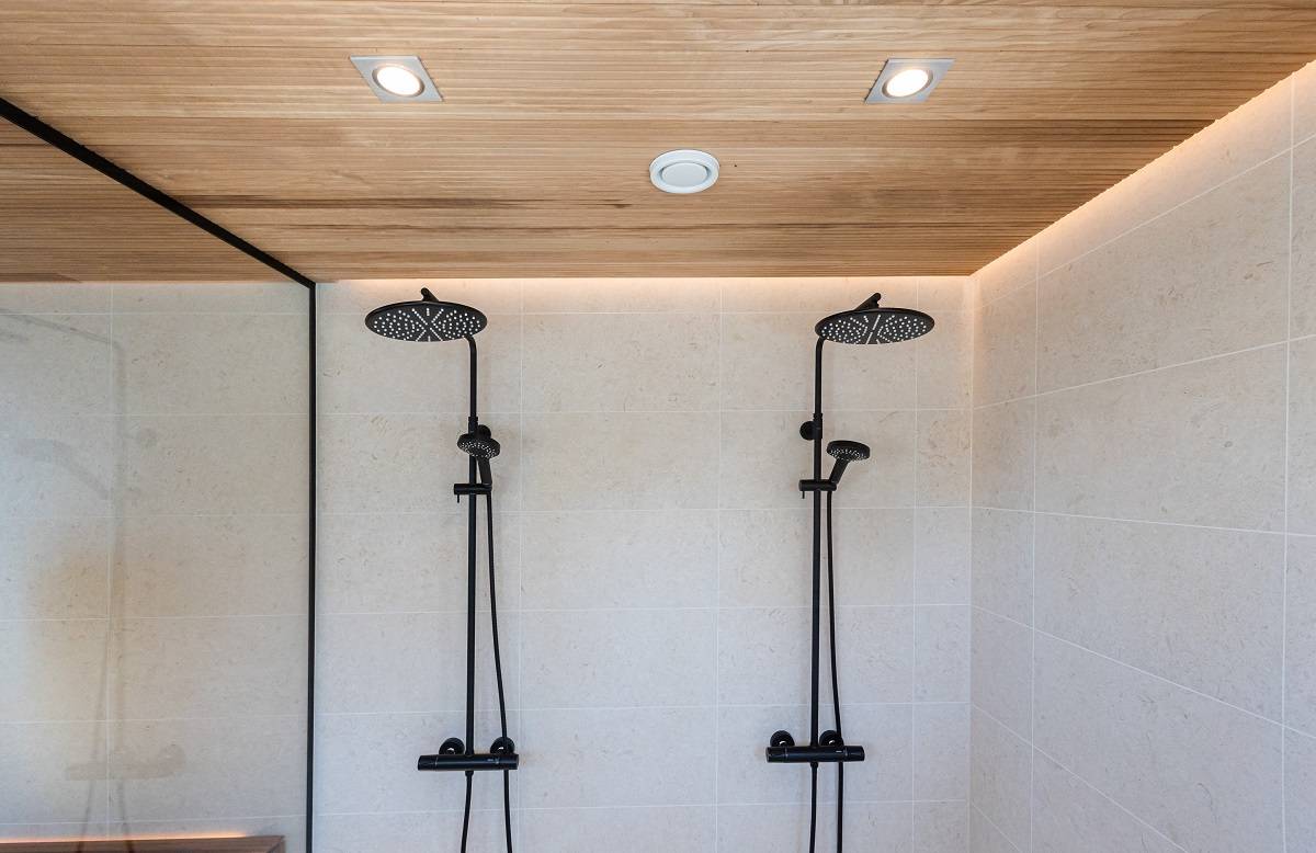 Das moderne Duschbad hat zwei schwarze Duschköpfe zu cremefarbenen Fliesen. Die holzgetäfelte Decke, die von LED-Einbauleuchten von LedStore.fi akzentuiert wird, sorgt für einen minimalistischen und eleganten Look. Der Duschbereich ist durch eine Glasaußenwand vom Rest des Raumes abgetrennt.