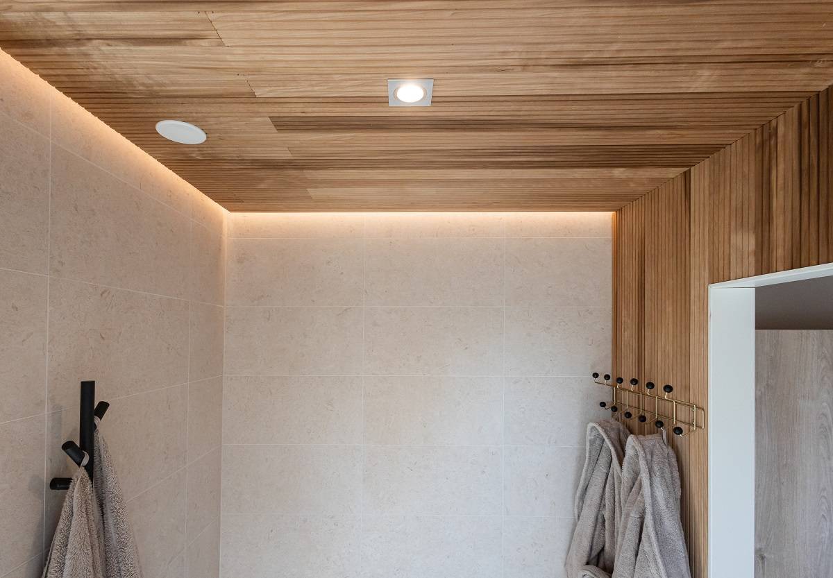 LedStore.fi bietet eine Lösung für das moderne Badezimmer mit moderner LED-Beleuchtung. Die beige gefliesten Wände, die Holzdecke und die eingelassene LED-Beleuchtung sorgen für einen frischen und modernen Look. Weiche graue Handtücher, die an schwarzen Haken aufgehängt sind, harmonieren mit der minimalistischen Einrichtung.  