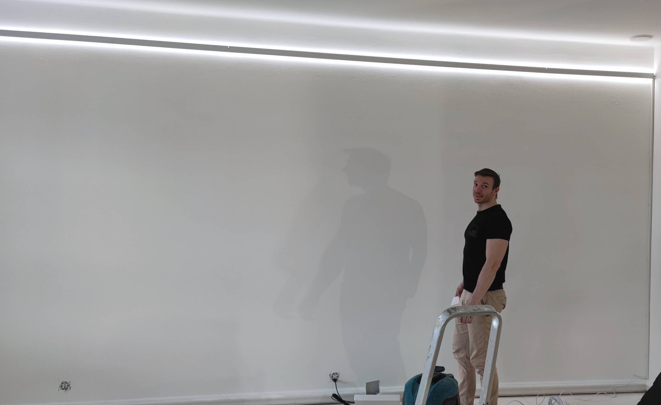 So installieren Sie indirekte LED-Beleuchtungsbänder Eine Person auf einer Leiter mit einem Werkzeug, in weicher LED-Beleuchtung von oben, vor einer weißen Wand. Auf dem Boden verstreute Kabel und Geräte in LED-Ladeneinrichtung.