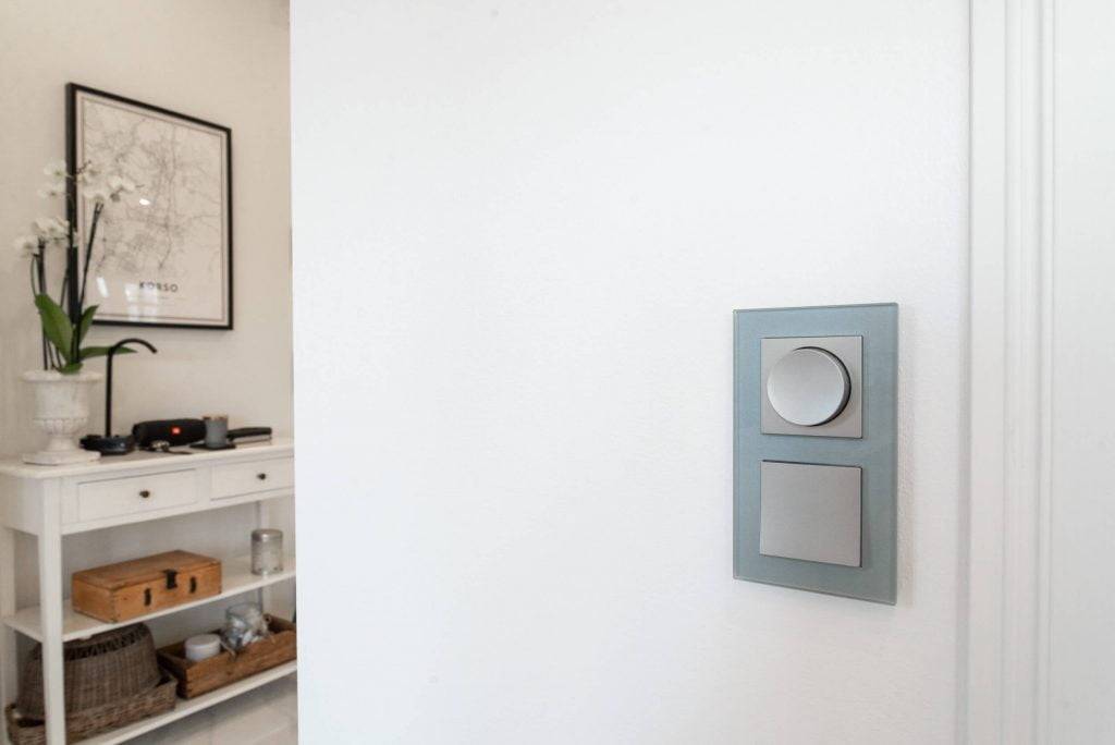 Moderner LED-Lichtschalter mit zwei Tasten an einer weißen Wand; weiße Konsole mit getöpferter Orchidee und gerahmter Karte in einem minimalistischen Raum.