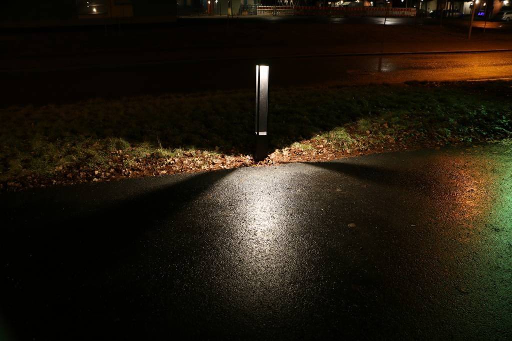 Eine LED-Straßenlaterne, die eine nasse Straße mit dreieckigen, von Gras gesäumten Mustern beleuchtet; eine städtische Kulisse mit weit entfernten Lichtern.