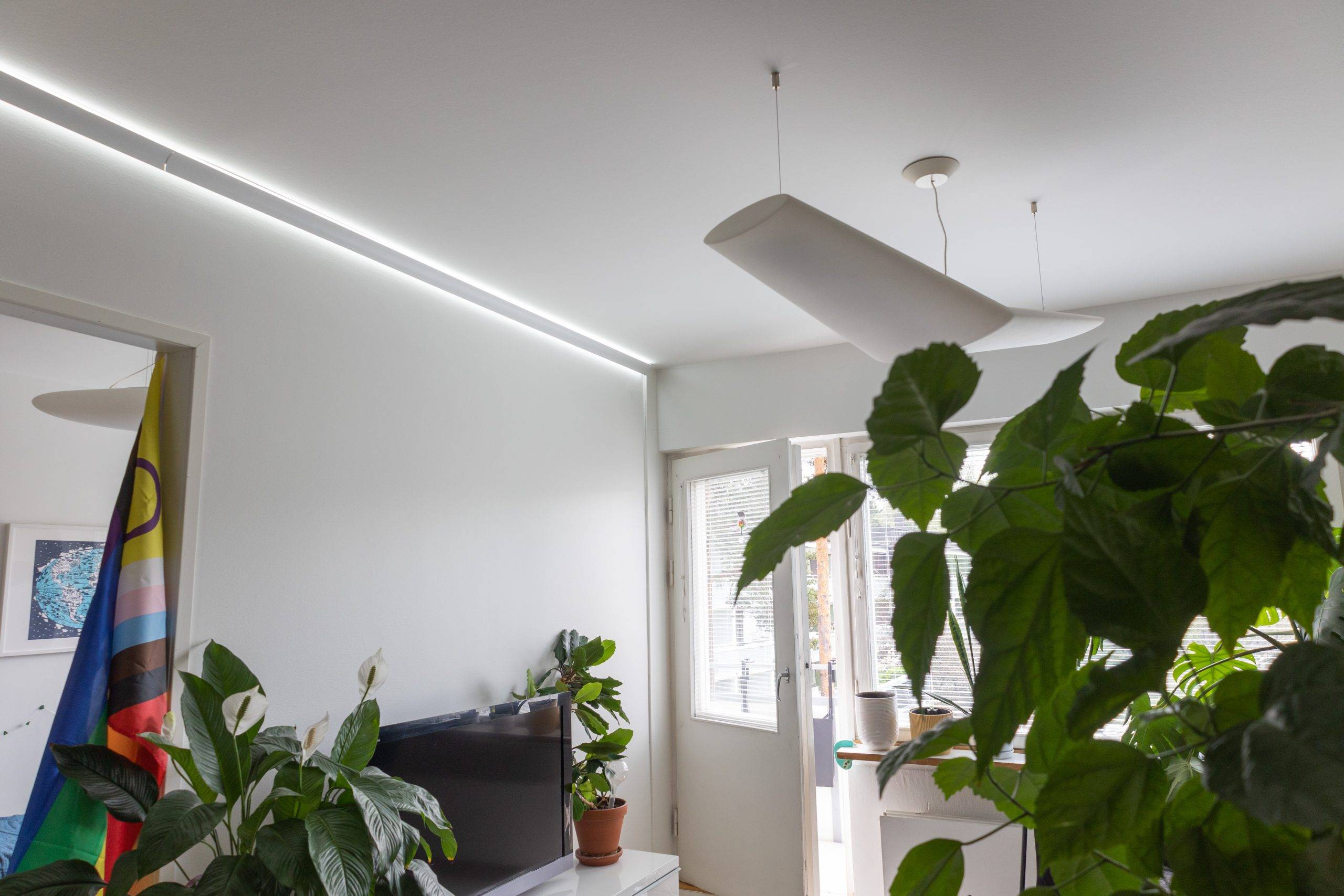 Ein modernes, minimalistisch gestaltetes Wohnzimmer mit großen Grünpflanzen, einem Fernseher und einer bunten Flagge. An der Decke befindet sich eine stilvolle LED-Leuchte neben eleganten LED-Elementen, während durch die Glastüren, die nach draußen führen, natürliches Licht einfällt.