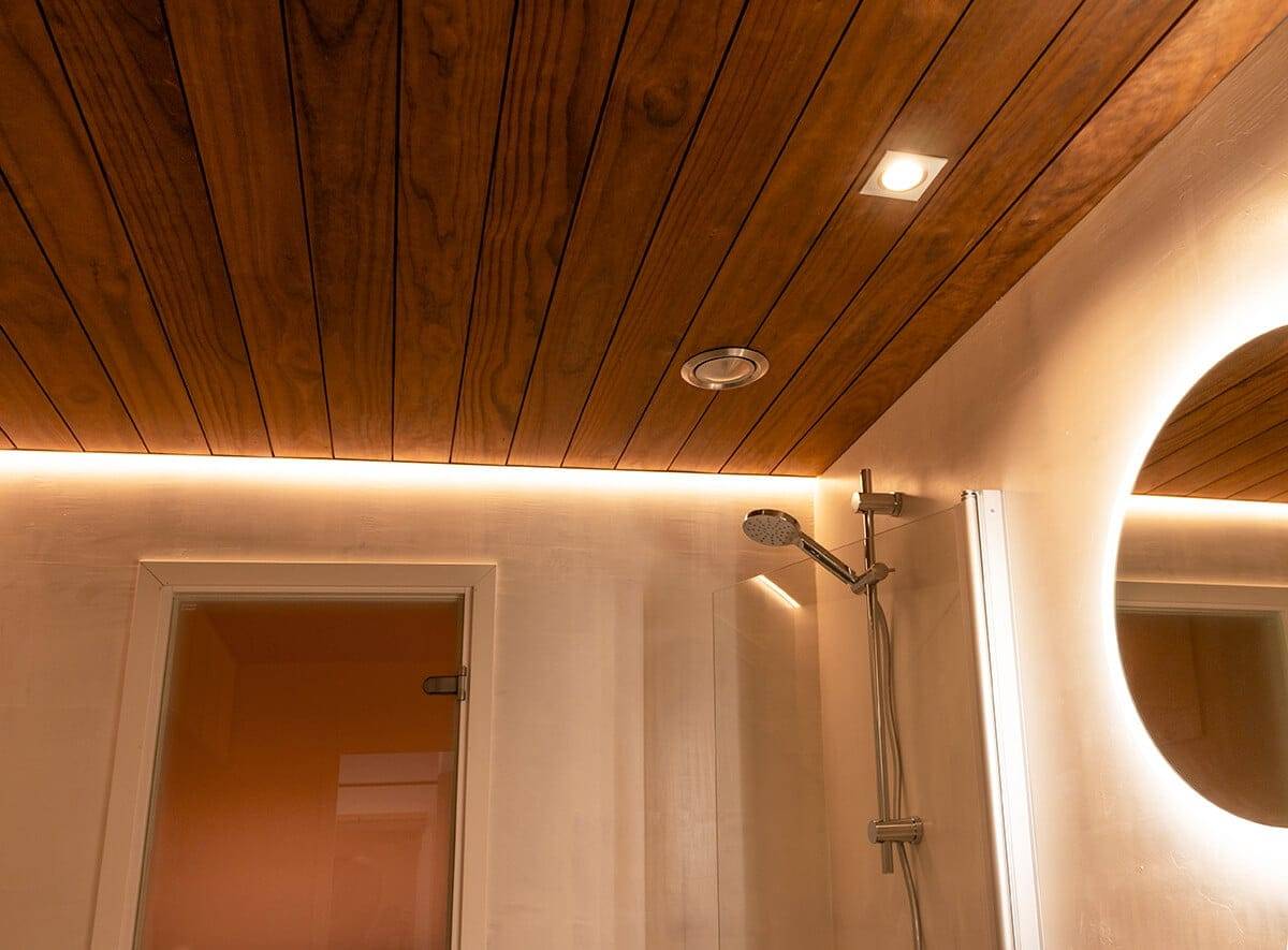 Badezimmer mit Holzdecke, LED-Umgebungsbeleuchtung, modernem Duschkopf, Scheinwerfer und rundem LED-Spiegel; Türöffnung links.