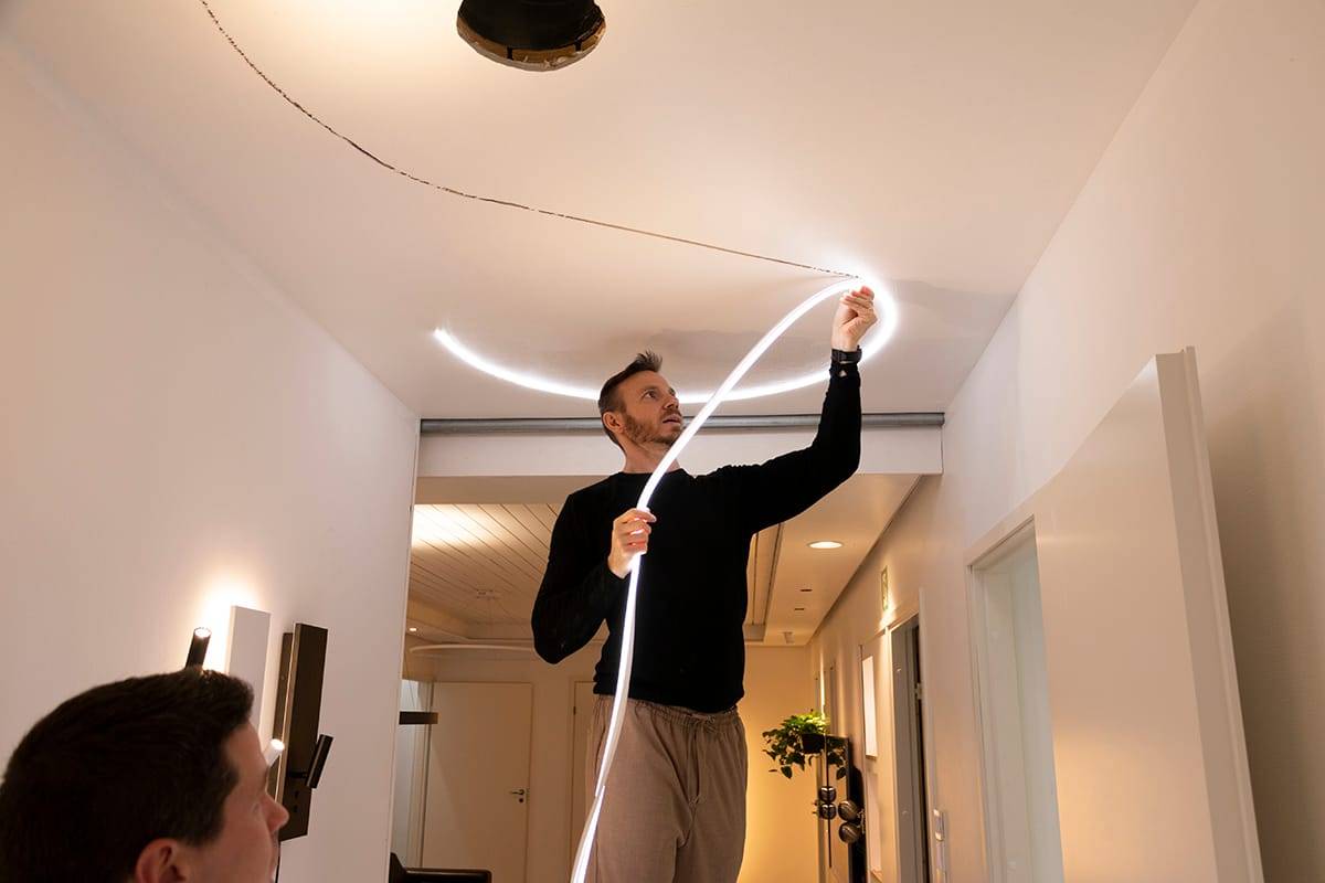 Eine Person bringt ein flexibles LED-Lichtband an einer modernen Decke an, wodurch die Beleuchtung im Raum verbessert wird. Schwarzer Pullover, beige Hose; eine weitere Person im Vordergrund, unscharf.