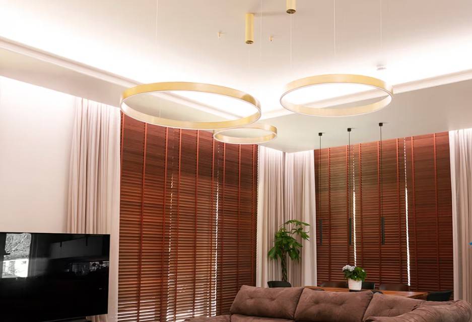 Das moderne Familienwohnzimmer in Kaitamäki, Espoo, verfügt über ein beeindruckendes Beleuchtungskonzept von LedStore.fi: große, goldfarbene Ringleuchten schaffen eine luxuriöse Atmosphäre zusammen mit passenden braunen Jalousien und einem gemütlichen Sofa. Abgerundet mit einem stilvollen Fernseher und üppigem Grün - das ist die Art von Beleuchtung, die Ihr Zuhause gemütlich und eindrucksvoll erscheinen lässt! 