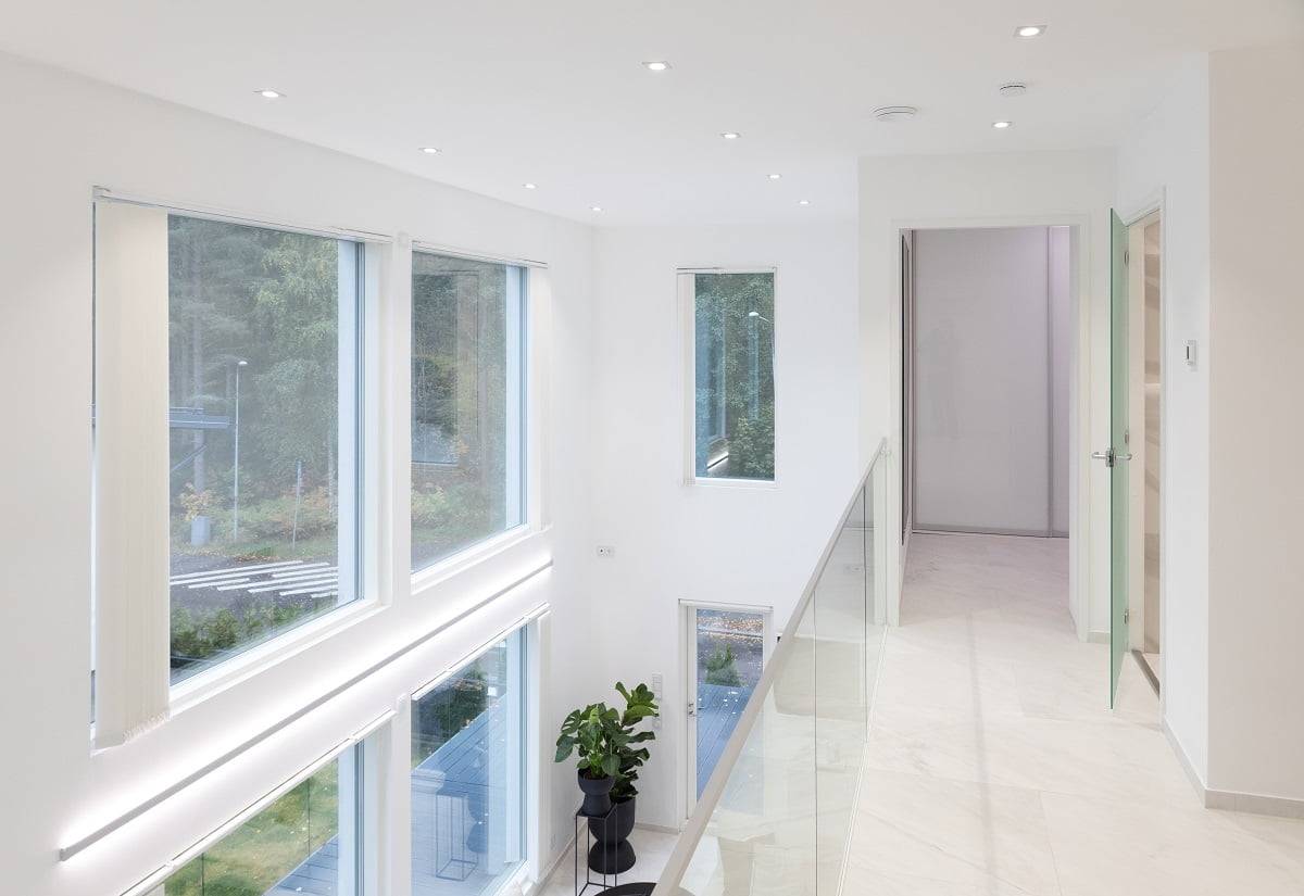 Ein heller und moderner Innenflur mit großen Fenstern auf beiden Seiten lässt natürliches Licht hereinströmen. Der Raum hat weiße Wände und eine LED-Einbauleuchte, die eine kinderfreundliche Atmosphäre schafft. Eine Glasbalustrade sorgt für Eleganz. Eine grüne Topfpflanze verleiht dem Ganzen einen frischen Farbklecks. Willkommen im hochwertigen LED-Beleuchtungssortiment von LedStore.fi und schaffen Sie den hellen Raum Ihrer Träume!