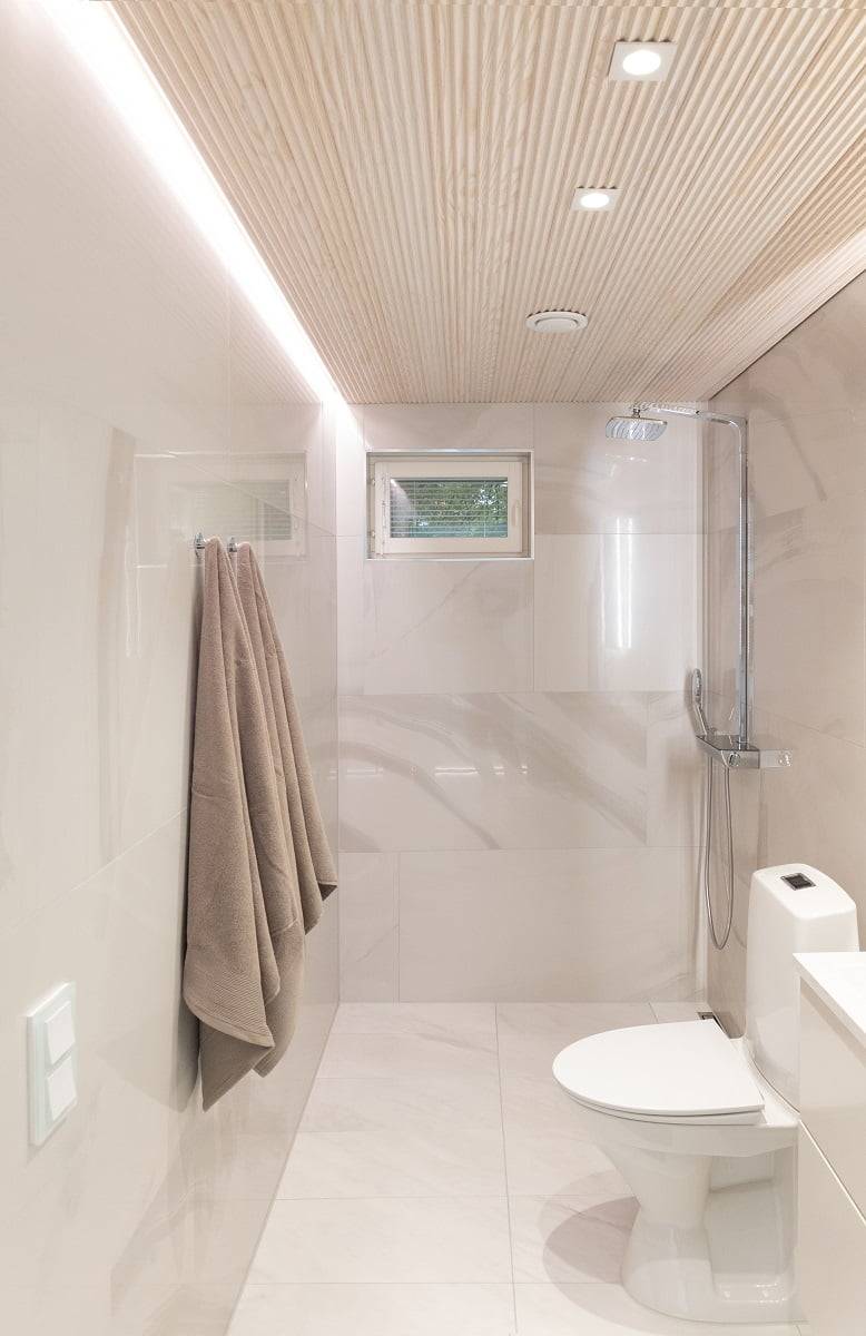 Ein modernes und minimalistisches Badezimmer mit hellbeigen Wänden und einer gefliesten Holzdecke. Ein an der Wand montierter Handtuchhalter mit zwei Handtüchern, ein kleines Fenster und ein durch eine Glaswand abgetrennter Duschbereich verleihen dem Raum Eleganz. Integrierte LED-Deckenleuchten sorgen für eine effektive Beleuchtung. LedStore.fi: Entdecken Sie LED-Beleuchtungslösungen, die perfekt für stilvolle und zeitlose Innenräume sind.   