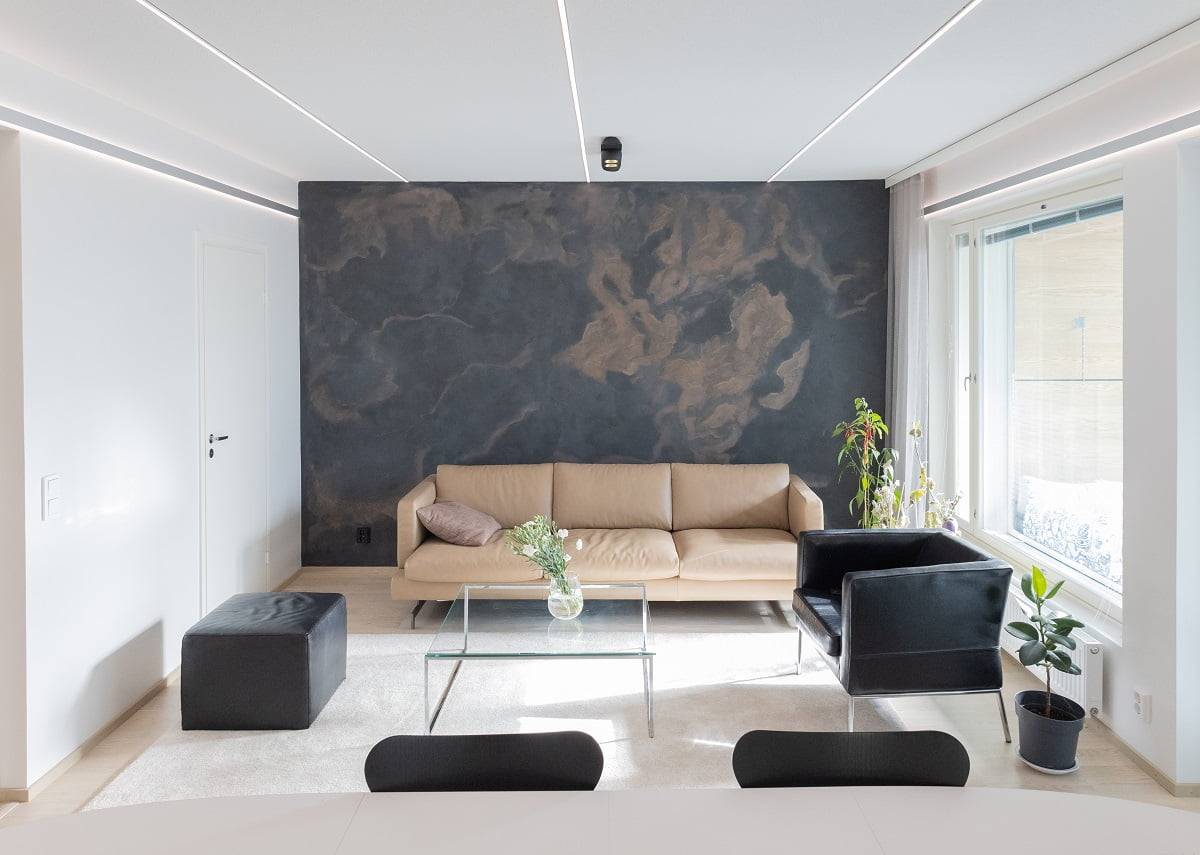Das moderne Wohnzimmer ist mit einem beigen Sofa, einem schwarzen Sessel und einem Couchtisch aus Glas eingerichtet, die zu der dunklen Wand passen. Neben dem Fenster steht eine Grünpflanze neben einem kleinen schwarzen Strohhalm. Bestellen Sie einen Strahler, ergänzt durch wilde LED-Leuchten von LedStore.fi.