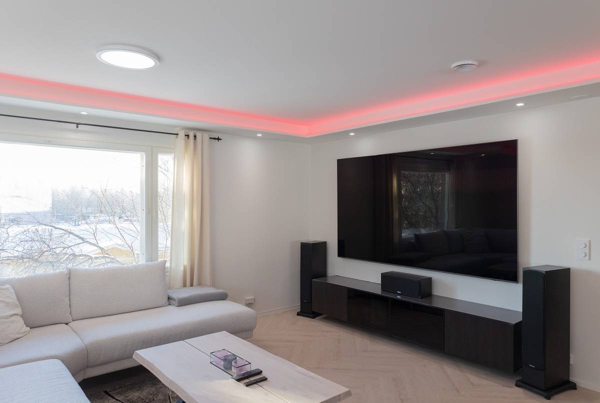 Ein modernes Wohnzimmer voller eleganter Atmosphäre. Ein großes Sofa lädt zum Entspannen ein, während ein heller Couchtisch aus Holz dem Raum Wärme verleiht. Ein an der Wand montierter Flachbildfernseher mit LED-Hintergrundbeleuchtung verleiht dem Raum einen modernen Touch. Verstärken Sie Ihr Erlebnis mit Bodenlautsprechern und genießen Sie die rote LED-Akzentbeleuchtung an der Decke. Ein großes Fenster lässt natürliches Licht herein, so dass der Raum großzügig und hell wirkt. All diese Elemente lassen sich mit den vielseitigen LED-Beleuchtungslösungen von LedStore.fi zu einer perfekten Umgebung kombinieren.
