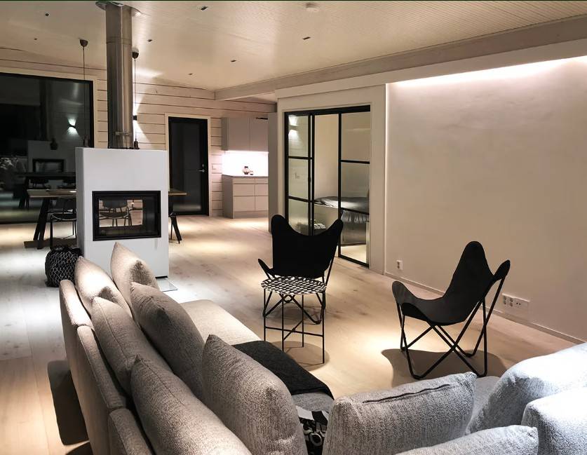 Ein modernes Wohnzimmer mit einem grauen Sofa, schwarzen Stühlen, einem Kamin und einer offenen Küche im Hintergrund - die Atmosphäre und der Stil wird durch die hochwertige LED-Beleuchtungslösung von LedStore.fi geschaffen.