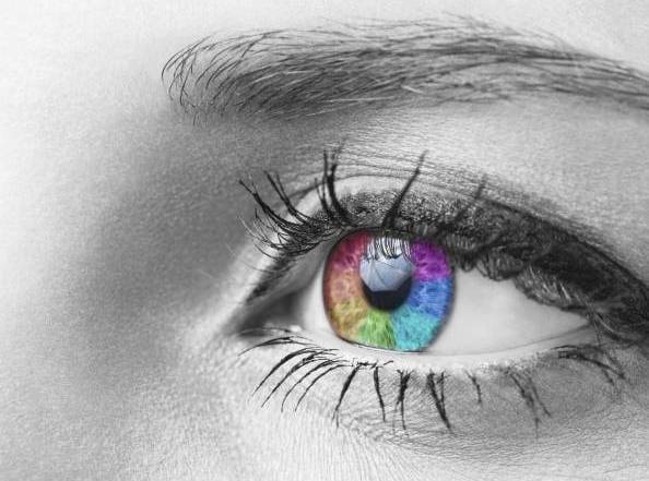 Grautonige Augen-Nahaufnahme mit LED-artig schillernder Iris in leuchtenden Farben: rot, blau, grün, violett; scharfer Kontrast zu monochromer Haut und Wimpern.