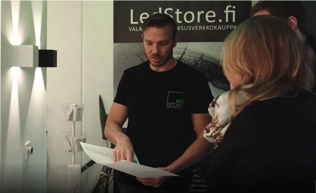 Ein Mann in einem schwarzen LED-Store-Shirt unterhält sich angeregt mit einer blonden Frau, eine weitere Person hört in der Nähe zu; ein "LedStore.fi"-Schild und helle LED-Lichter im Hintergrund.
