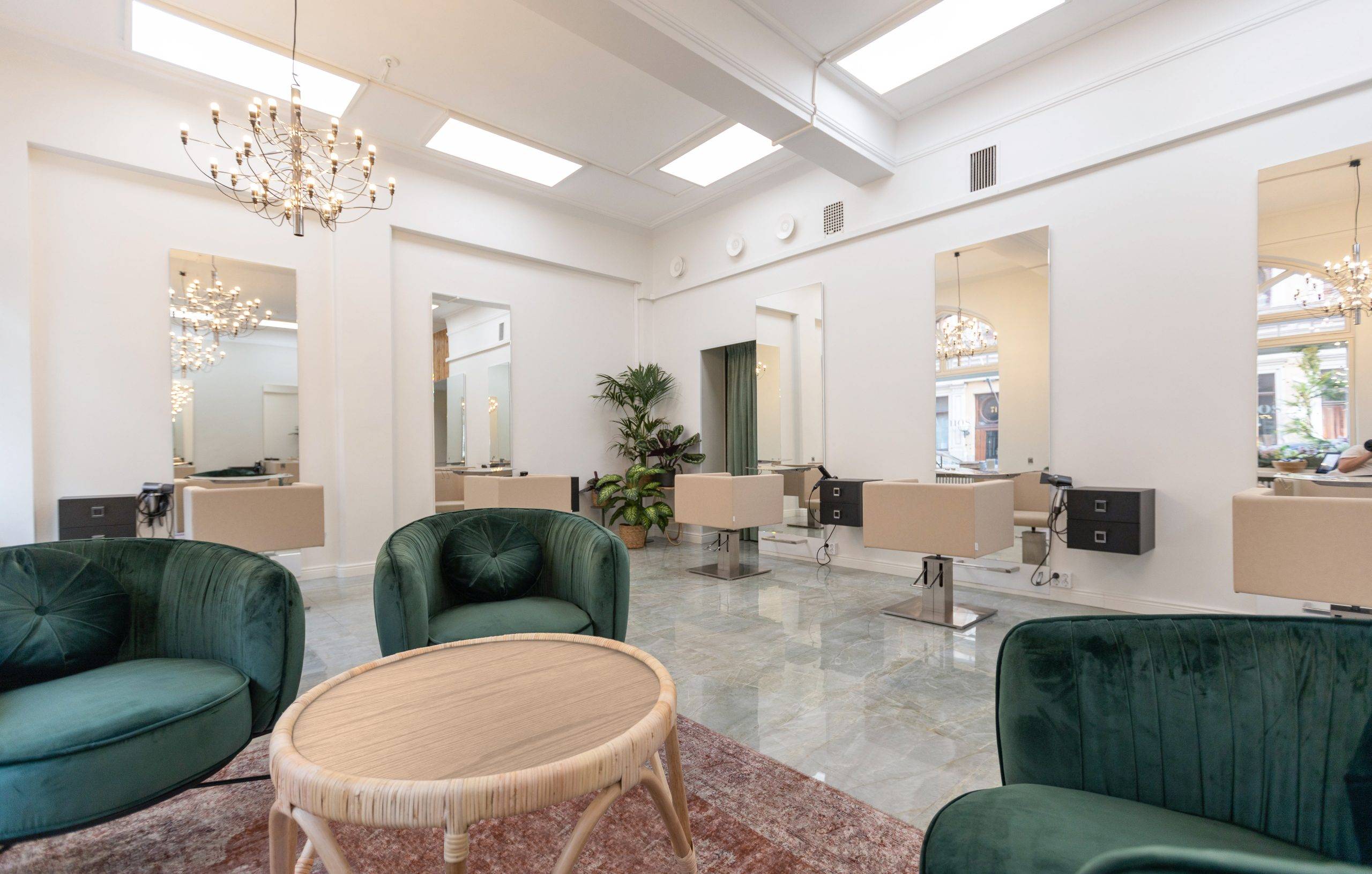 Ein modernes und helles Saloninterieur mit grünen Samtstühlen um einen runden Tisch mit rotem Teppich. Salonarbeitsplätze mit Spiegeln und beigen Stühlen säumen die Wände. Sie werden von an der Decke montierten LED-Kronleuchtern beleuchtet. Große Fenster lassen natürliches Licht herein, während Grünpflanzen das Ambiente des Raumes verstärken. In dieser Art von Umgebung sind die hochwertigen LED-Beleuchtungslösungen von LedStore.fi ideal, denn sie sorgen für ein perfektes Gleichgewicht zwischen Licht und Energieeinsparung, ohne dass die Eleganz darunter leidet.