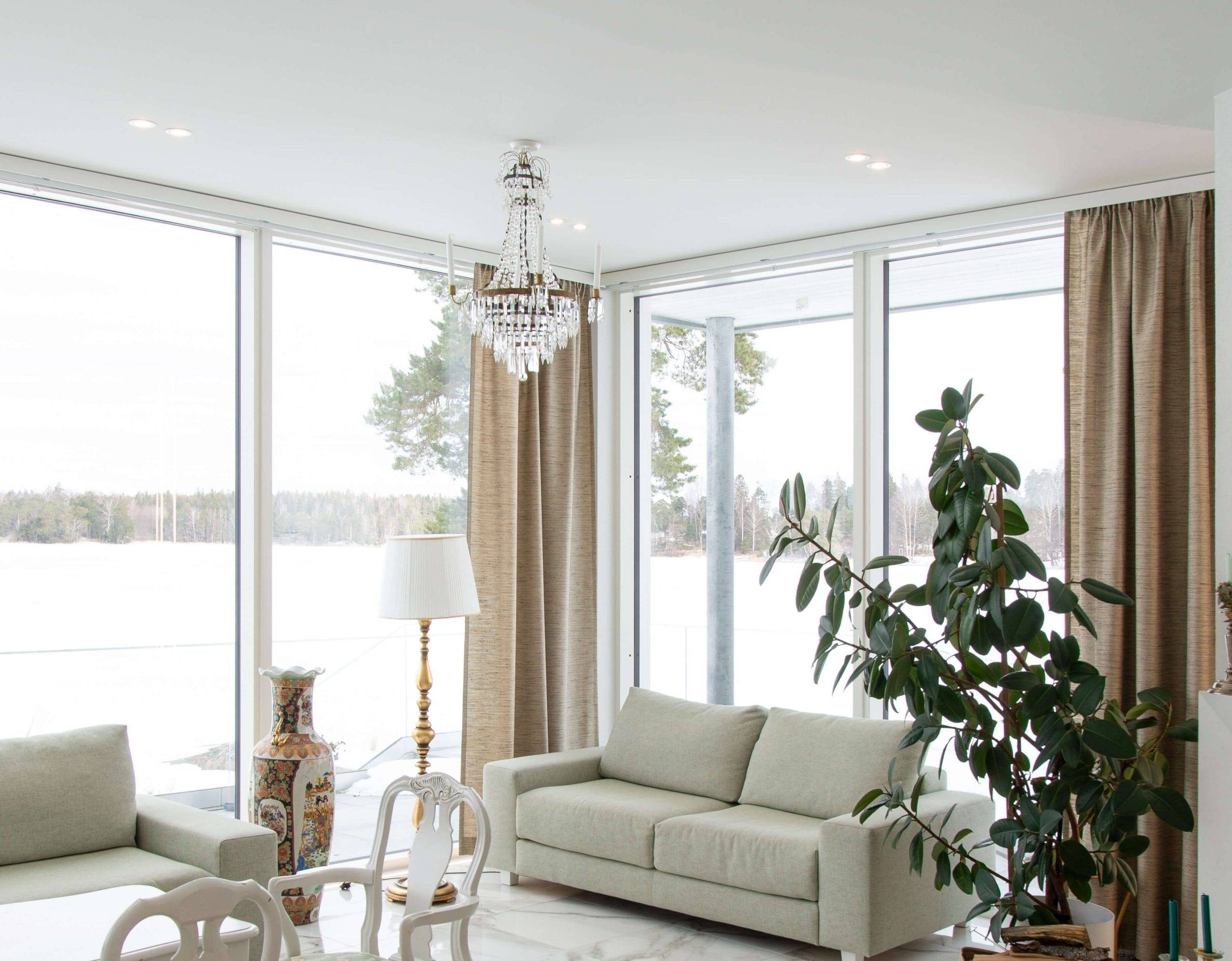 Das helle Wohnzimmer mit großen Fenstern und Blick auf die verschneite Landschaft verfügt über ein weißes Sofa, eine große Grünpflanze und einen stilvollen Kronleuchter mit LED-Leuchten, die die Schönheit des Raumes unterstreichen. Beige Vorhänge ergänzen eine dekorative Vase und eine Stehlampe, während im Hintergrund ein Esstisch mit weißen Stühlen glänzt. Schauen Sie sich die große Auswahl an LED-Leuchten bei LedStore.fi an und schaffen Sie eine ebenso beeindruckende Beleuchtungslösung für Ihr Zuhause!