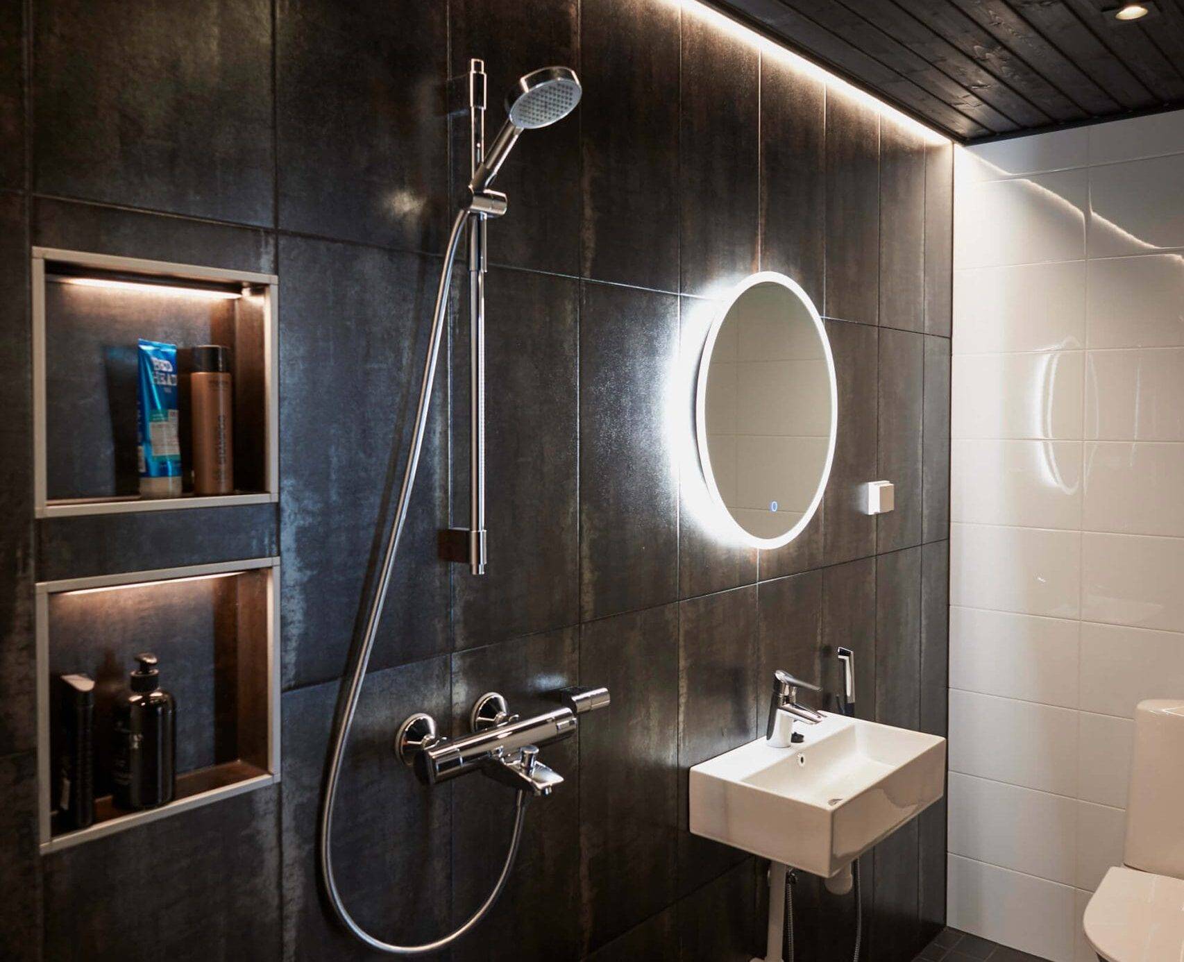 Stilvolles Badezimmer mit dunklen Fliesen, LED-beleuchtetem Spiegel über dem Waschbecken, Dusche, eingebauten Regalen und schwarzer Decke. Eine moderne Toilette vervollständigt den stilvollen Look.