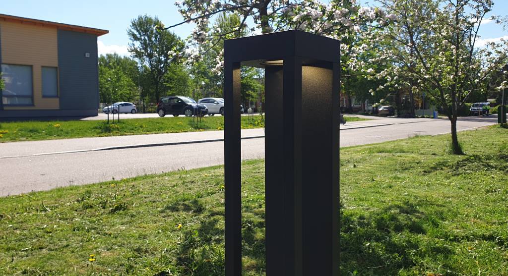Ein moderner schwarzer Laternenpfahl mit LED-Licht entlang der Straße; blühende Bäume, geparkte Autos und ein Gebäude mit großen Fenstern im Hintergrund unter einem sonnigen Himmel.