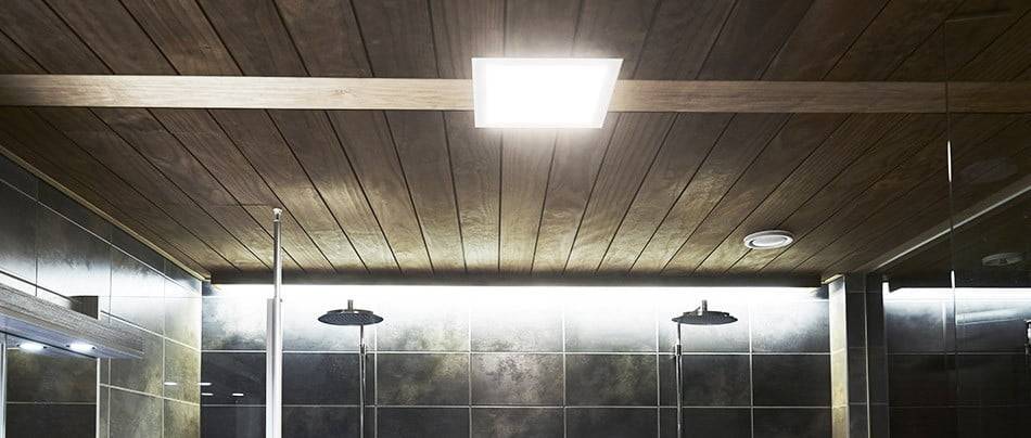 Modernes Badezimmer mit Holzdecke, quadratischer LED-Leuchte, dunkel gefliesten Wänden, zwei Regenduschköpfen und einer warmen Atmosphäre mit sanfter LED-Beleuchtung.