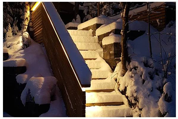 Eine schneebedeckte Treppe in der Abenddämmerung, mit einem warmen Licht, das vom Geländer auf die Treppe scheint.  