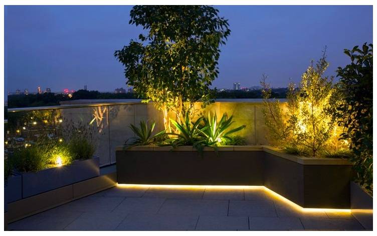 Erhöhtes Blumenbeet, das von unten mit einem warmfarbenen LED-Streifen beleuchtet wird. Das Blumenbeet ist außerdem mit weiteren warmtonigen Spotlights ausgestattet.  